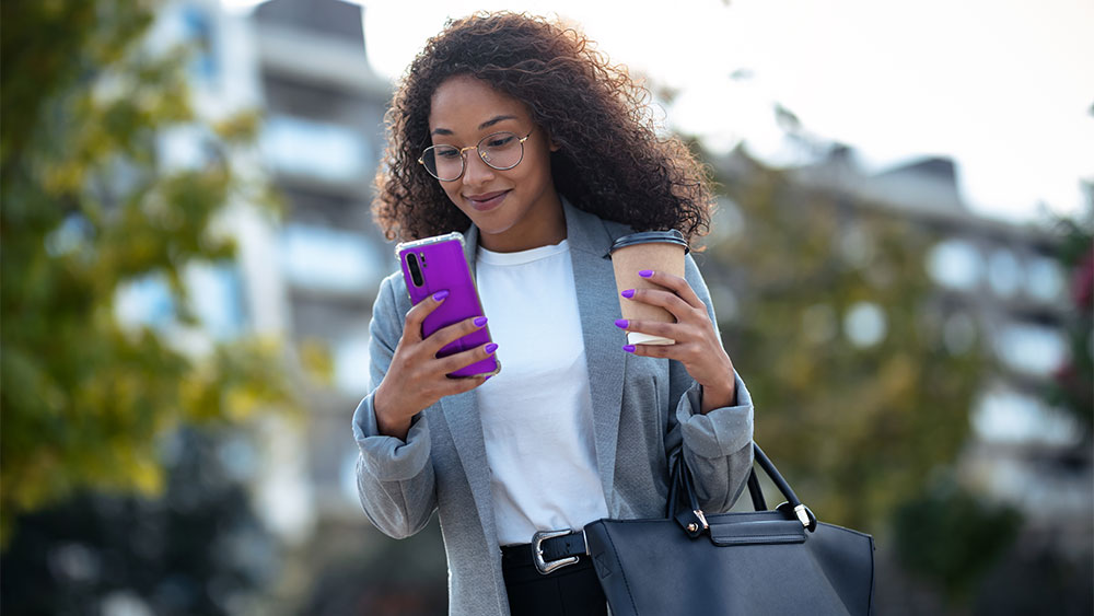 Mulher sorridente utilizando um dos melhores celulares de 2025 enquanto caminha ao ar livre com um café na mão.