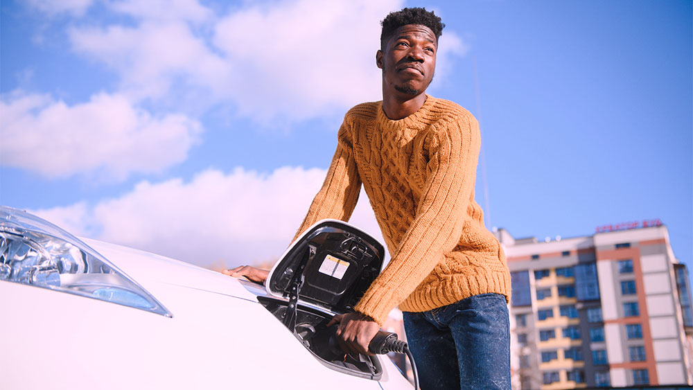 Pessoa conectando cabo de carregamento em automóvel branco, simbolizando o uso de veículos elétricos e a mobilidade sustentável.