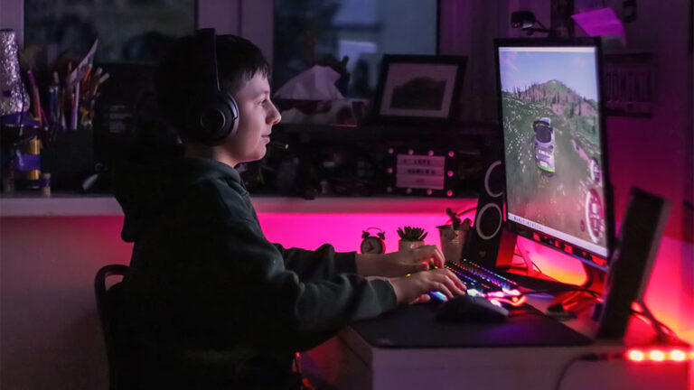 Jovem utilizando um Setup Gamer com monitor, teclado mecânico iluminado e headset, em ambiente com luzes LED coloridas.